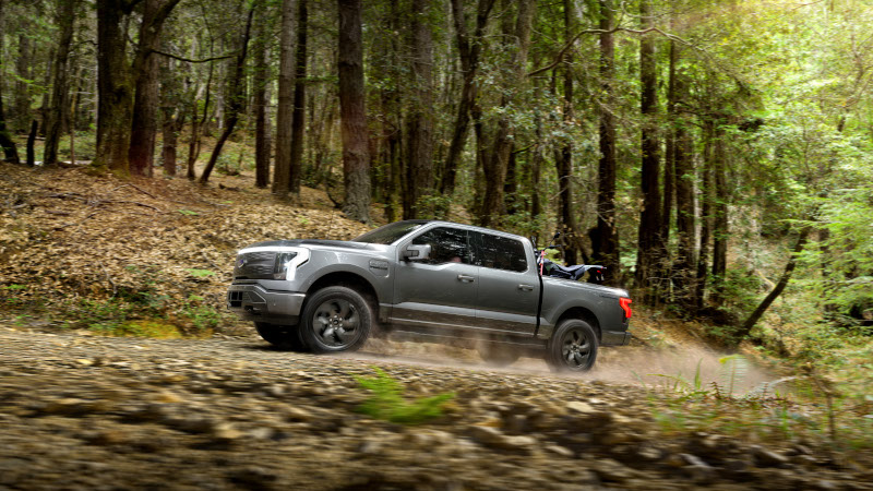 Pickup Truck Adventure in the Wilderness Silver pickup truck with dirt bike in bed driving on a forest trail, kicking up dust among towering trees.